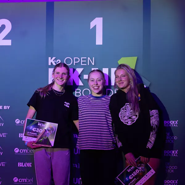 Drie jonge vrouwen staan glimlachend voor een groene podiumachtergrond waarop de cijfers 1 en 2 te zien zijn. Twee vrouwen houden certificaten vast; de vrouw in het midden staat op de hoogste trede van het podium. De verlichting is paars en blauw.